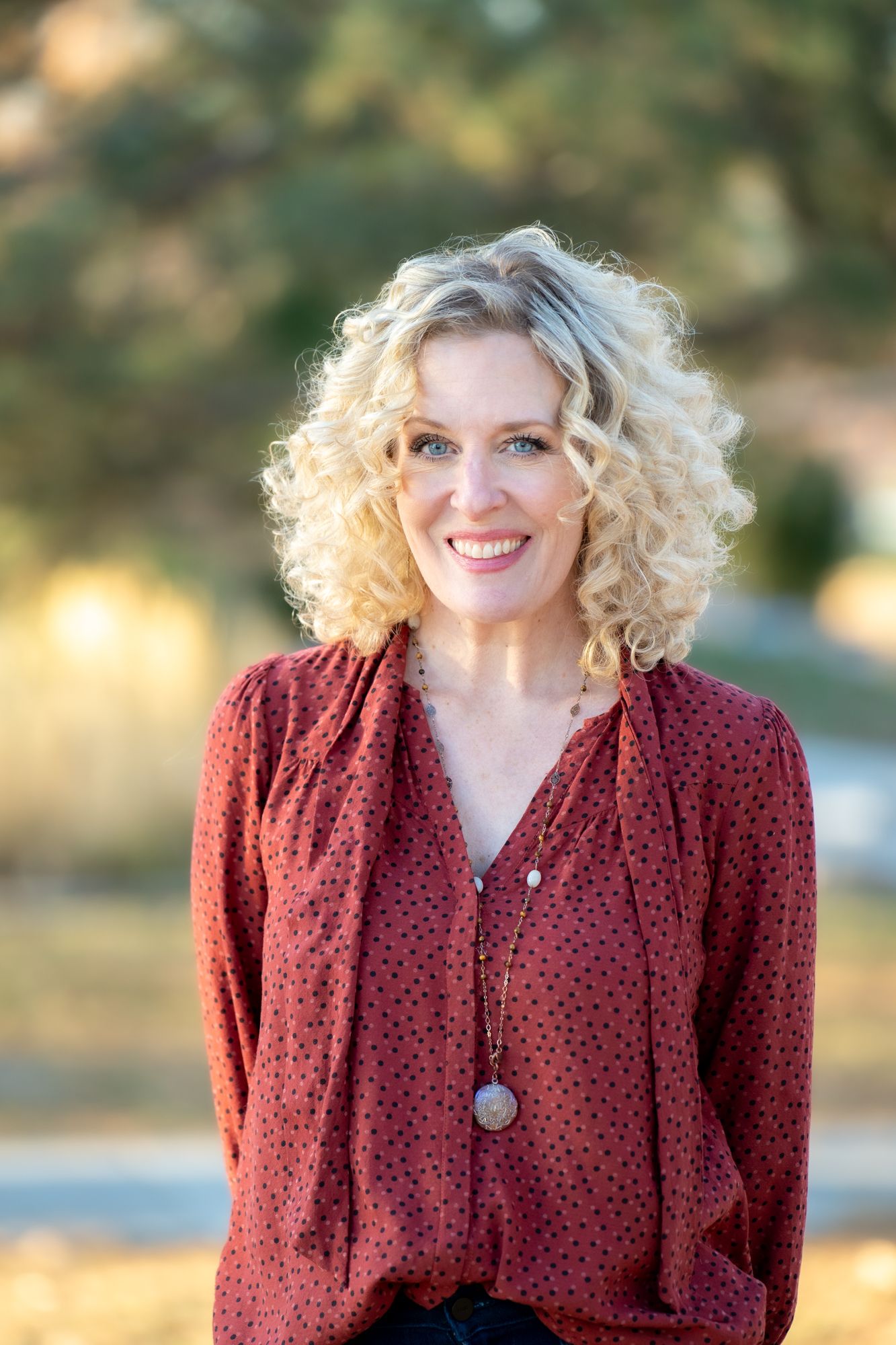 Professional portrait of woman with blonde curly hair wearing burgundy blouse and pendant necklace outdoors.