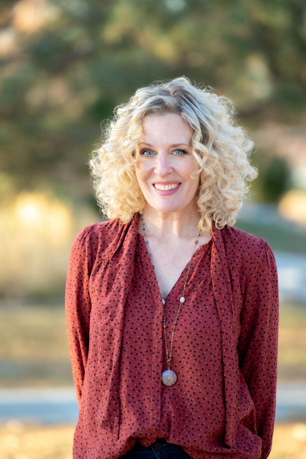 Professional portrait of woman with blonde curly hair wearing burgundy blouse and pendant necklace outdoors.