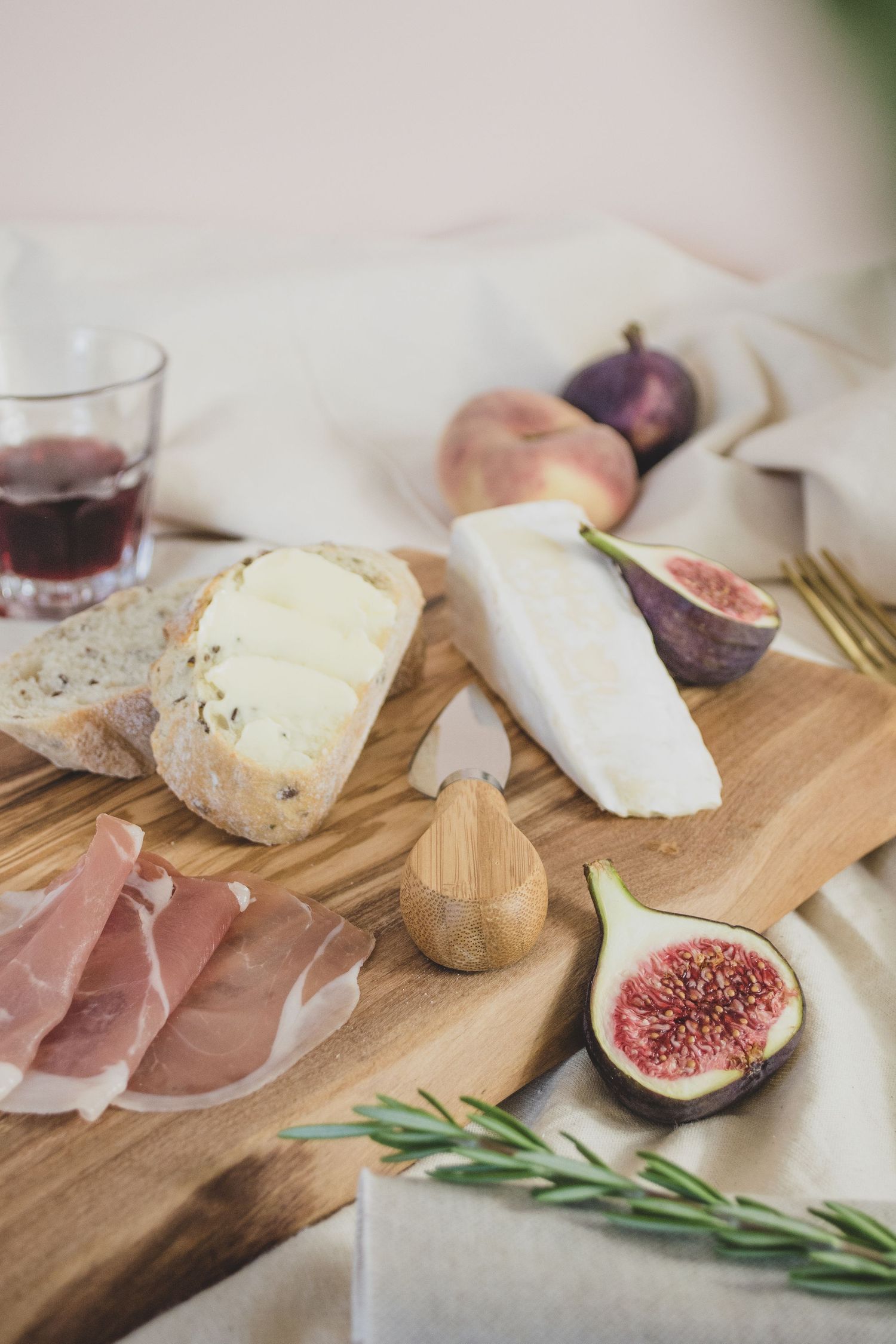 lifestyle photo of wooden board with bread, meats, cheese and wine with fruit on a neutral backdrop