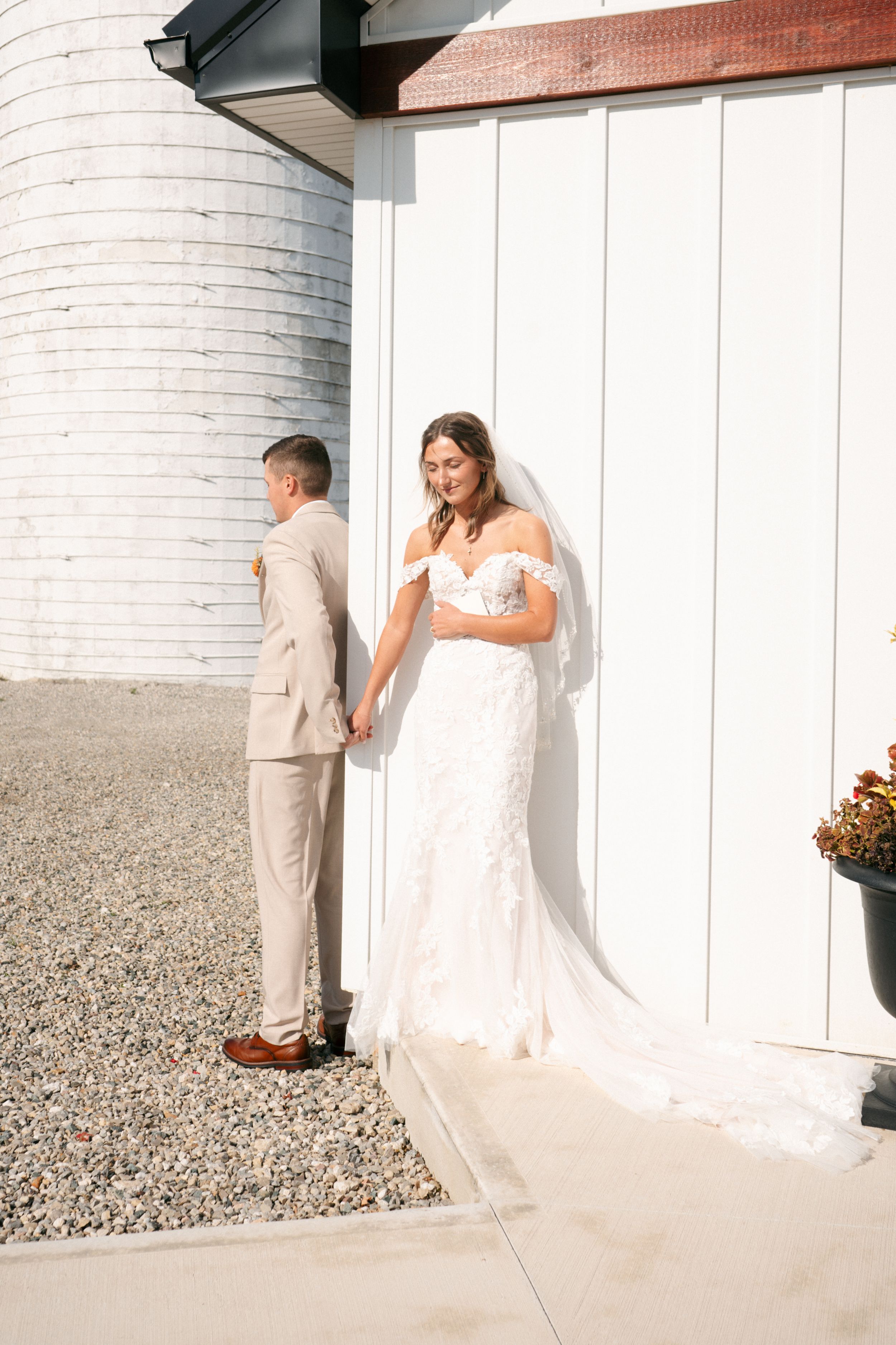 Bride in lace off-shoulder gown holds grooms hand outside white barn venue on sunny wedding day.
