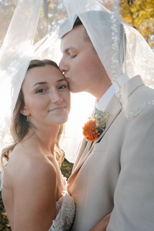 Groom kisses bride on cheek under flowing veil with orange boutonniere in fall sunlight.