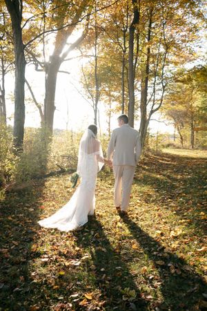 Bride and groom walk hand in hand through sunlit autumn forest with golden foliage.