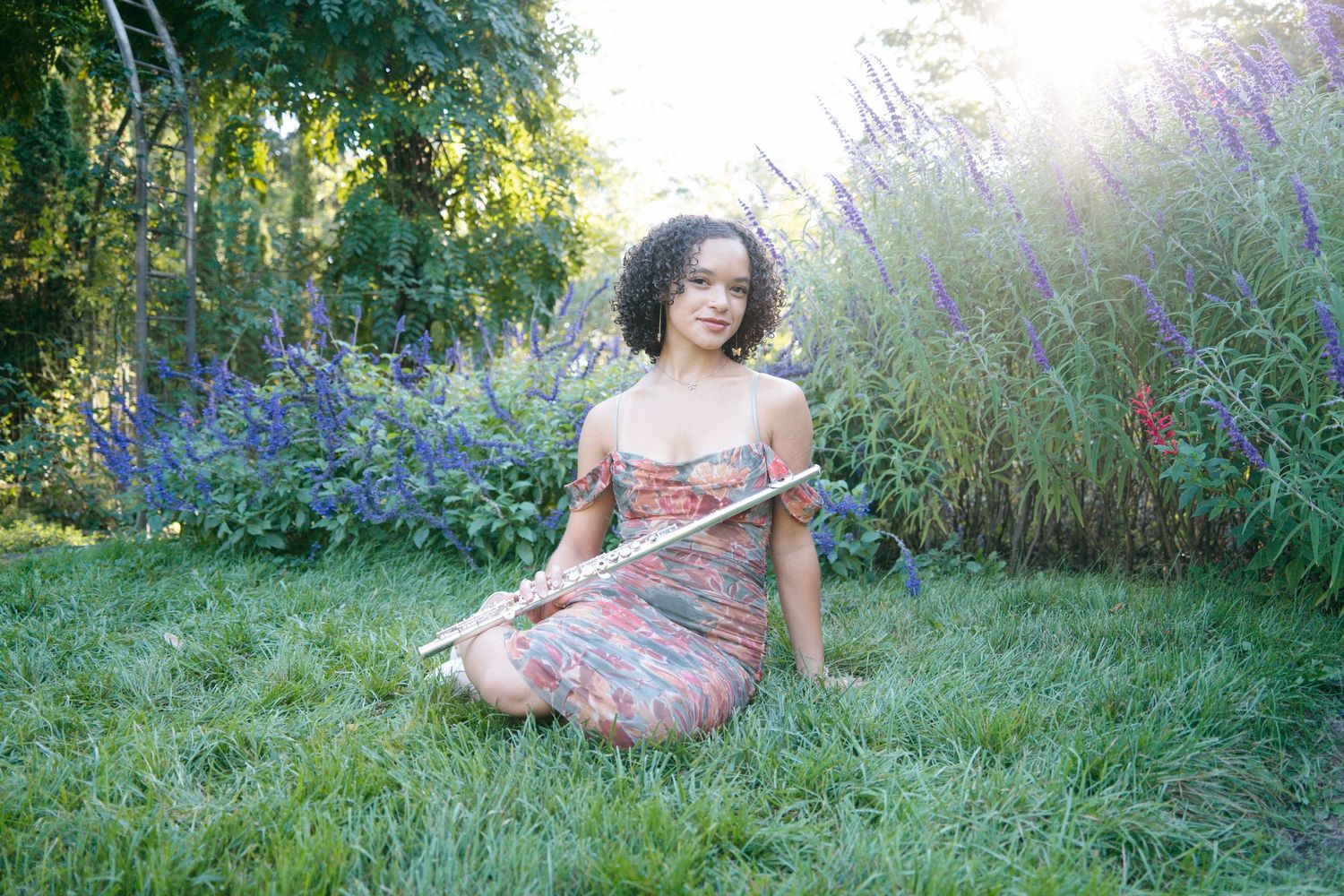 Young woman in floral dress sits cross-legged on grass holding a flute in a sunny garden.