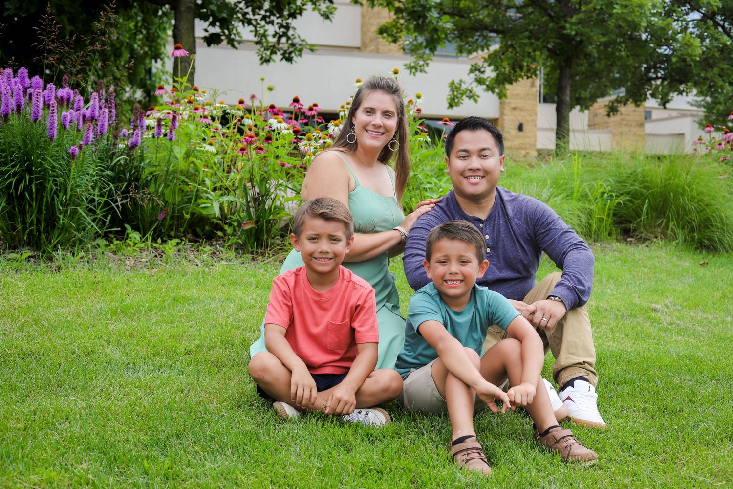 A family group sits together on grass with purple flowers blooming in the garden background.
