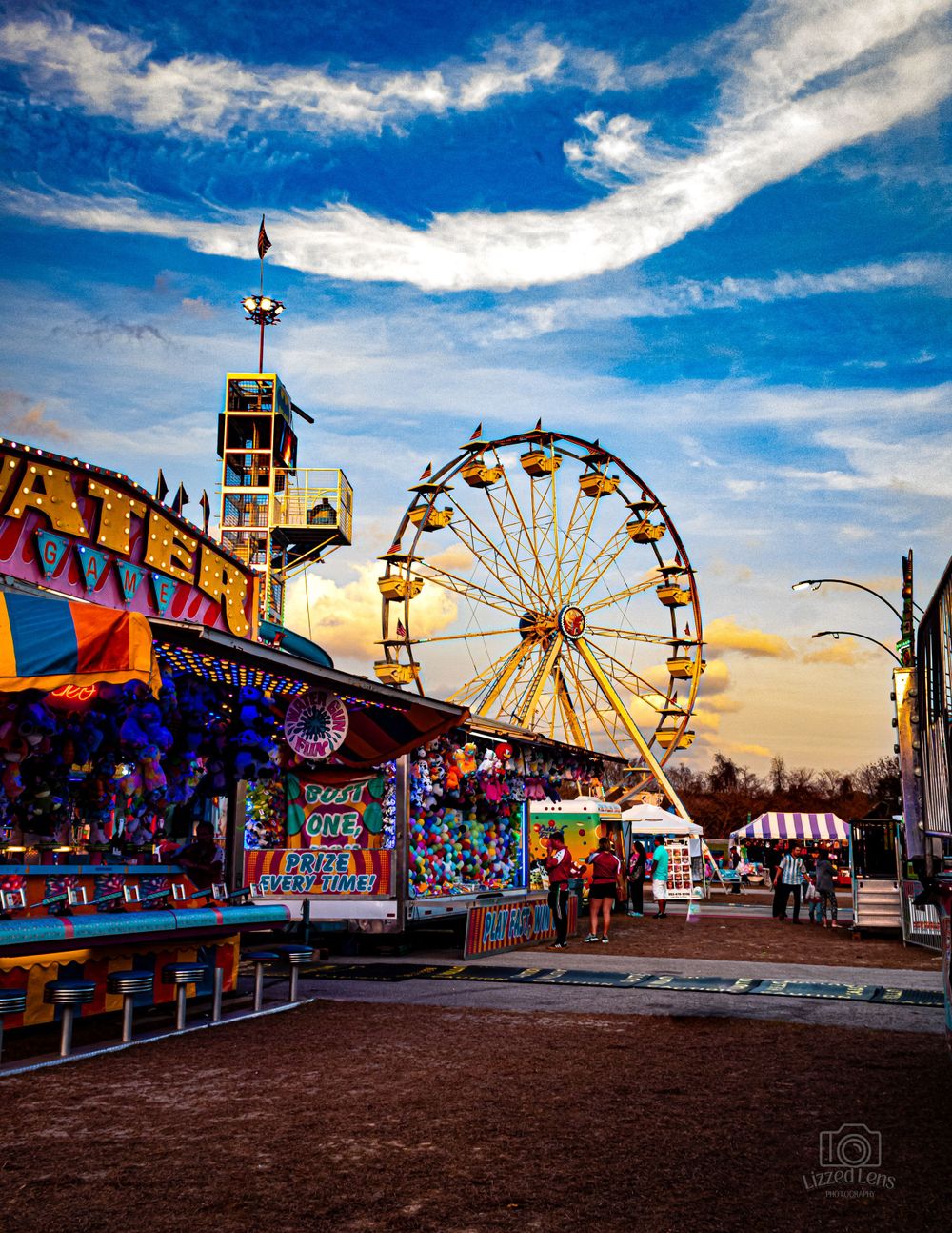 A colorful carnival midway with a Ferris wheel and game booths at sunset.