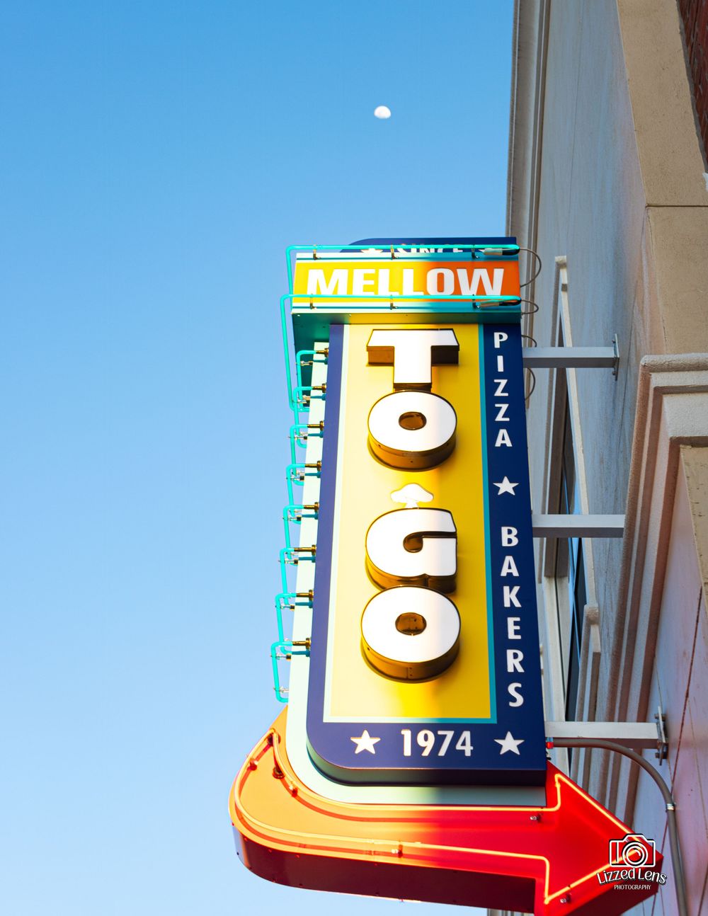A retro Mellow To Go Pizza Bakers neon sign from 1974 against a bright blue sky.