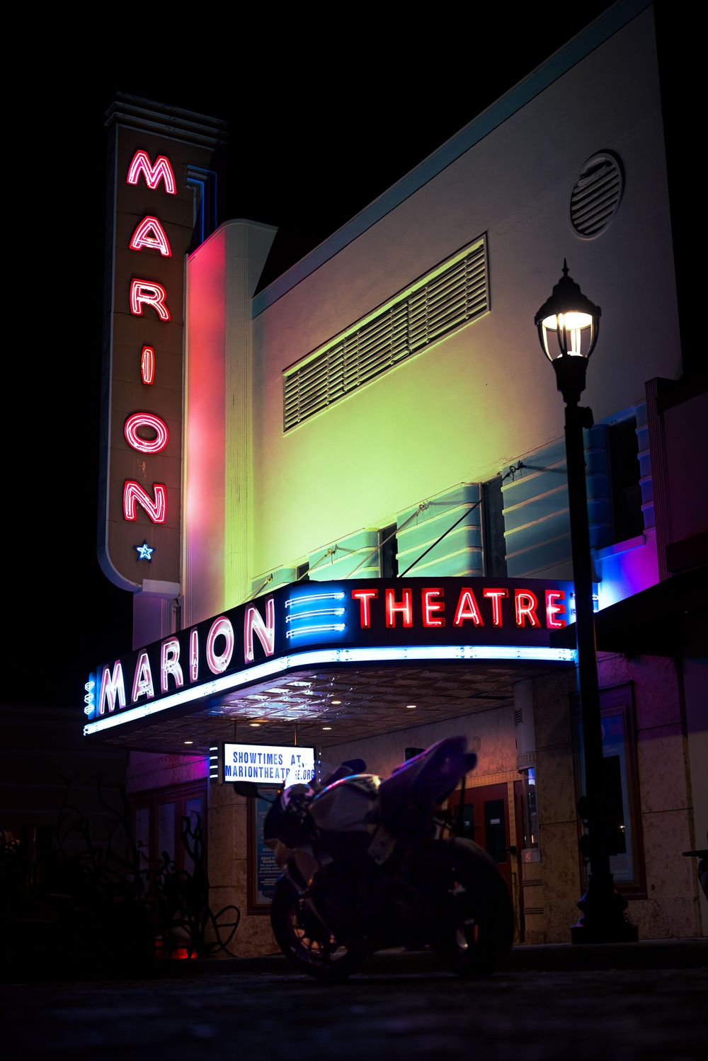 Neon-lit Marion Theatre marquee glowing in vibrant colors at night with a street lamp and motorcycle in foreground.