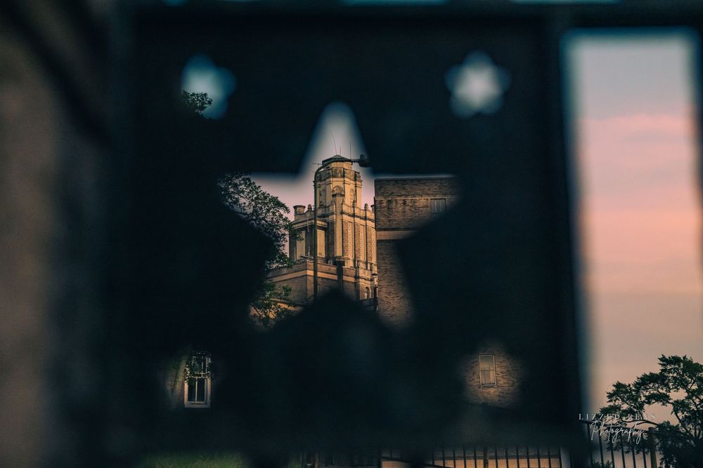 Historic stone tower building viewed through a decorative iron gate with bokeh light effects at dusk.
