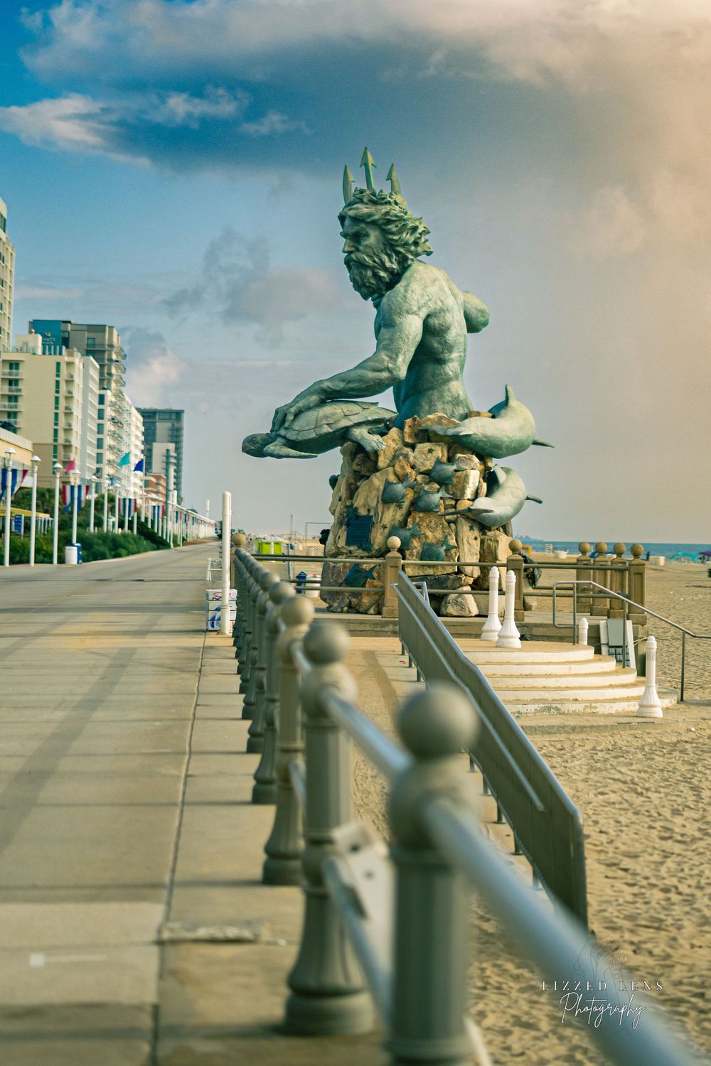 King Neptune bronze statue on Virginia Beach boardwalk with ocean and city skyline behind.