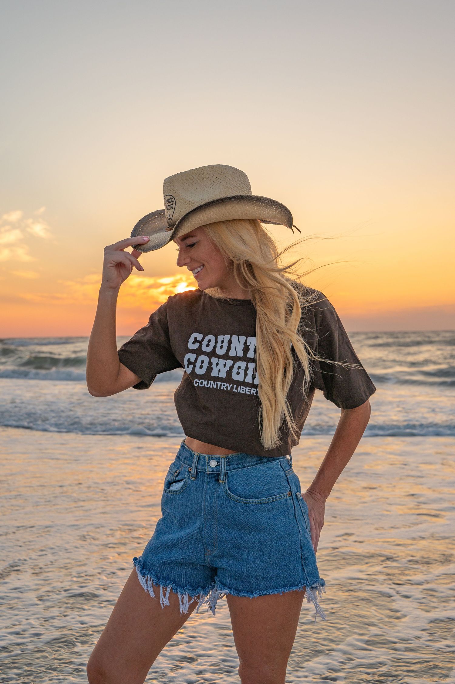 Person in cowboy hat and denim shorts poses on beach at sunset wearing country themed shirt.