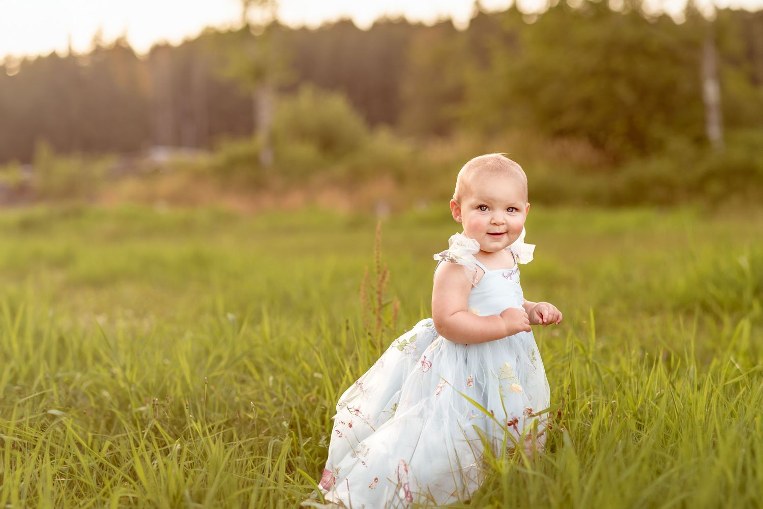A toddler in a light blue dress smiles while walking through a summer meadow at sunset.