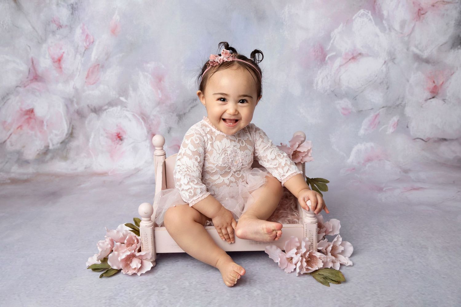 Baby in lace dress sits among pink floral decorations on light backdrop.