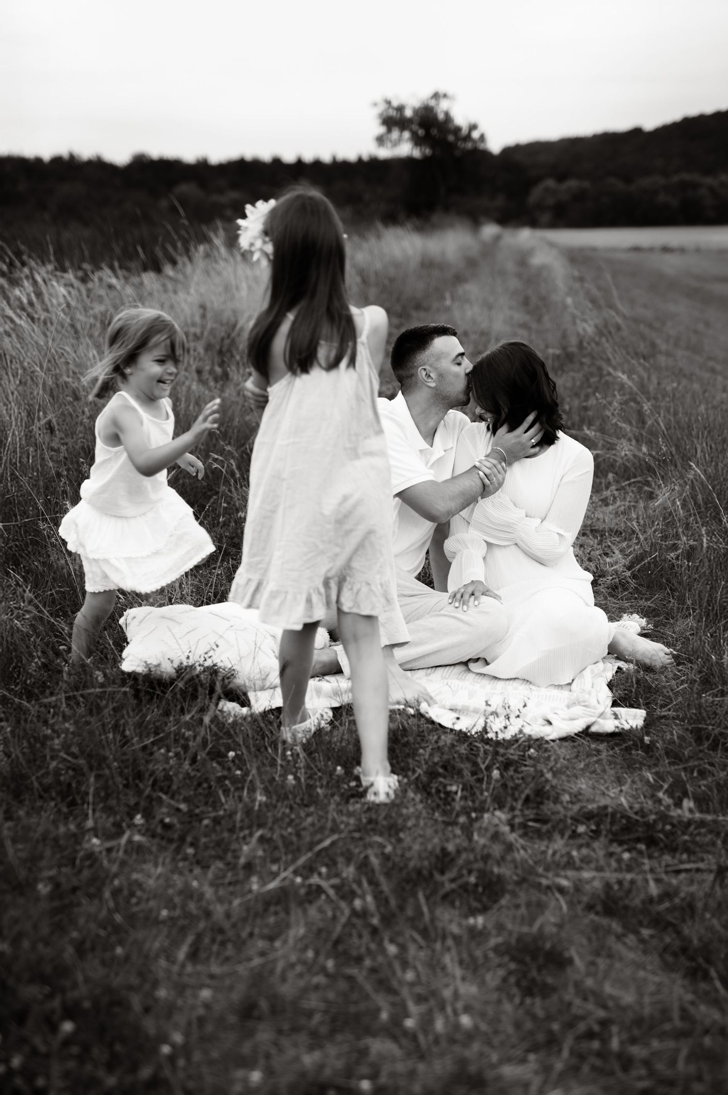 A baby in denim overalls shares a small cake smash moment with parents while sitting on a white blanket.