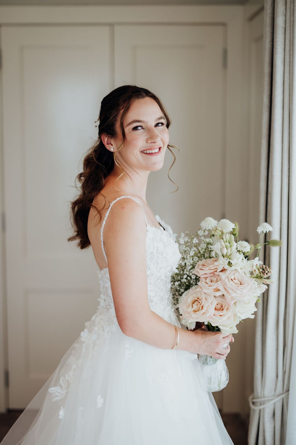 Bride in white ballgown holding floral bouquet poses by window.