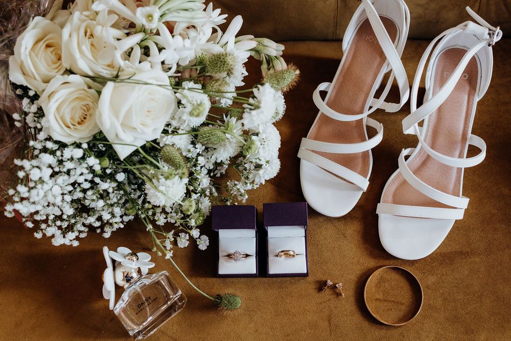 Wedding details flat lay with white roses bouquet, strappy sandals, rings and perfume bottle.