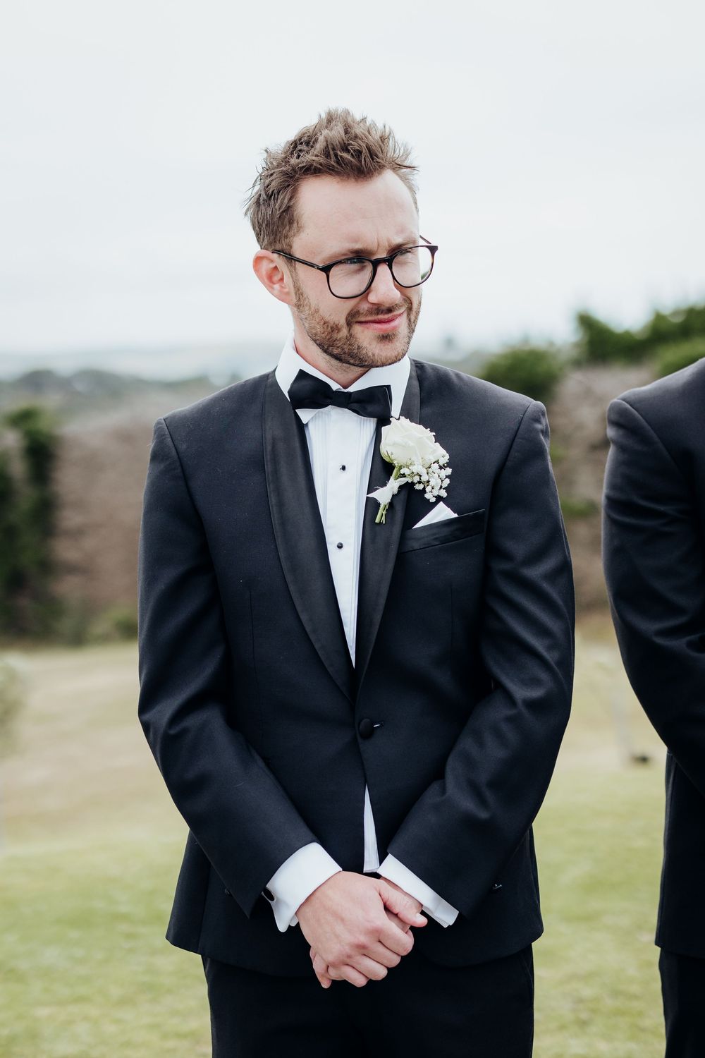 A groom in a classic black tuxedo and bowtie smiles during an outdoor ceremony.