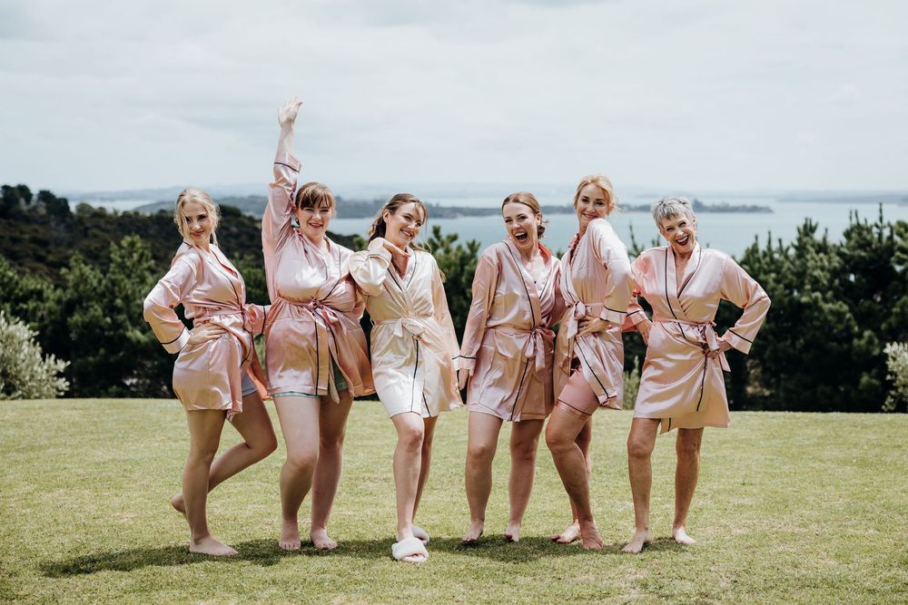 Bridesmaids in blush pink dresses pose playfully on a grassy outdoor venue with ocean views.
