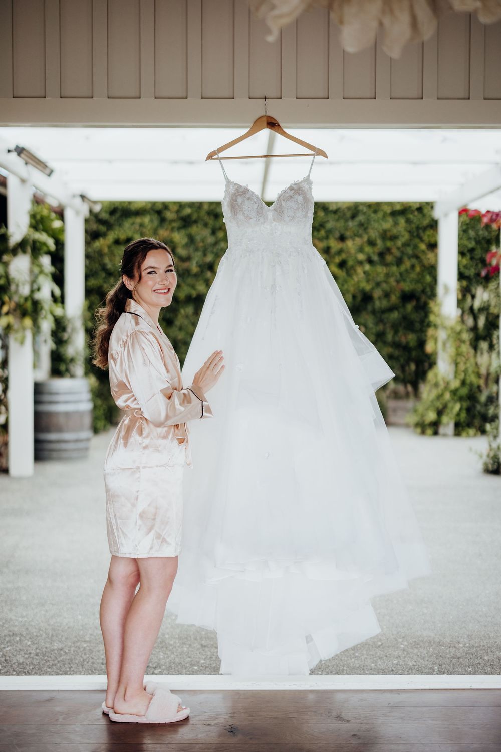 Small girl in a blush pink robe stands next to flowing white wedding dress during wedding preparations.