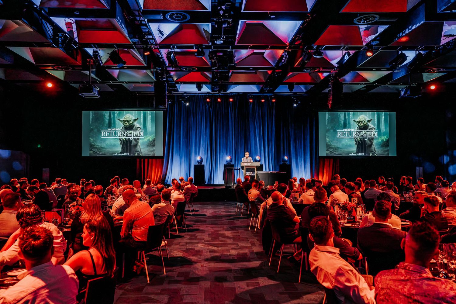 Large conference venue with stage and dual projection screens bathed in red lighting while attendees sit at round tables.