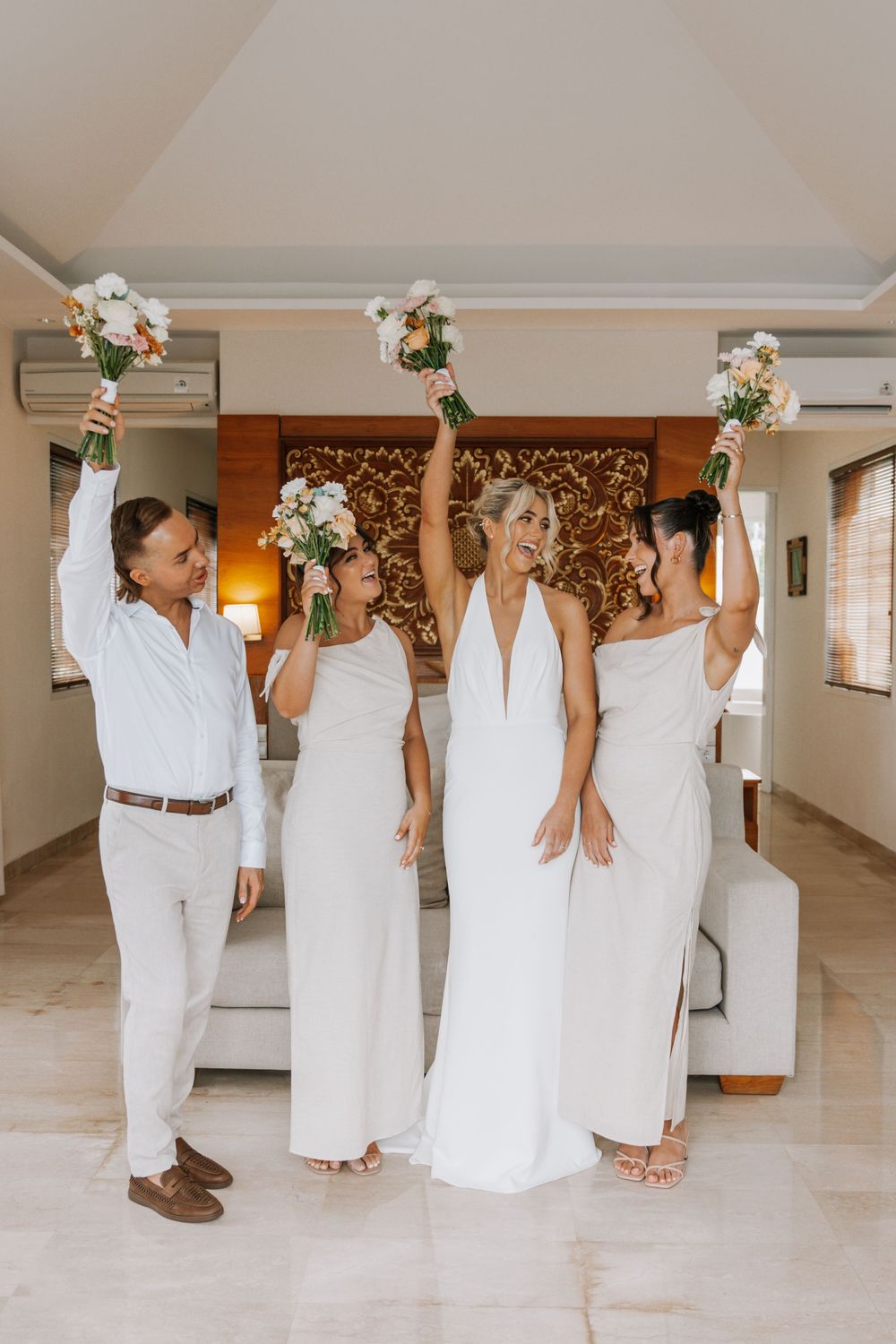 Wedding party members in white attire hold up champagne bottles in celebration along a light-colored hallway.