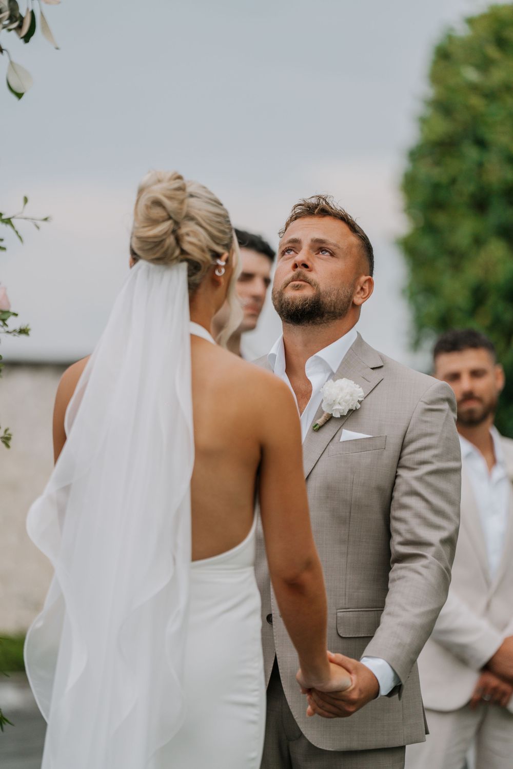 A groom in a light gray suit gazes intently during an outdoor wedding ceremony.