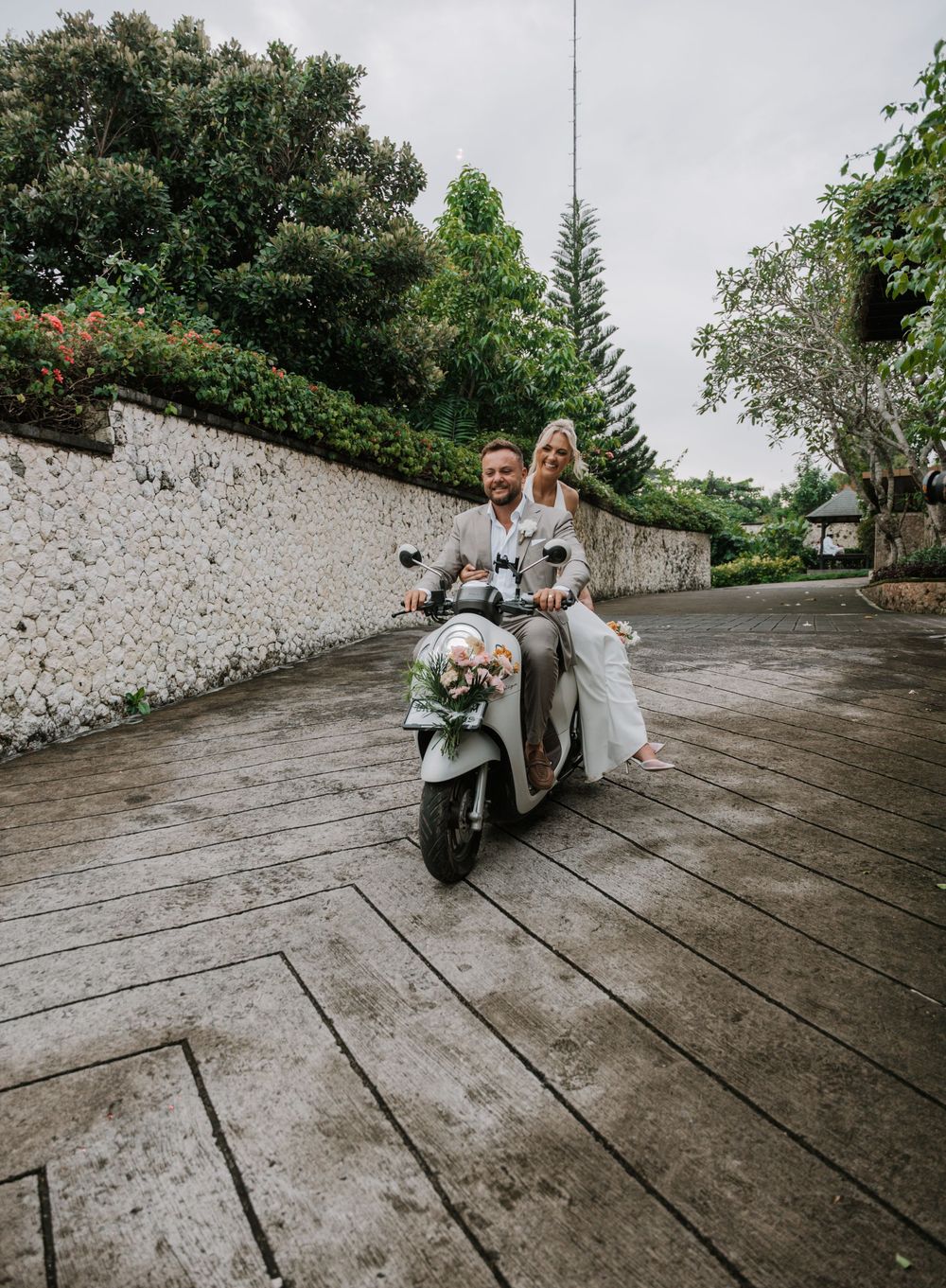 A couple riding together on a white scooter along a stone-walled path surrounded by lush greenery on a cloudy day.