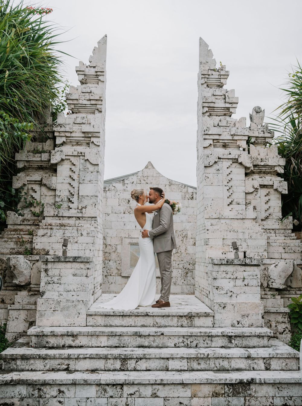 Couple in wedding attire share romantic moments on white stone steps with tropical foliage in the background.