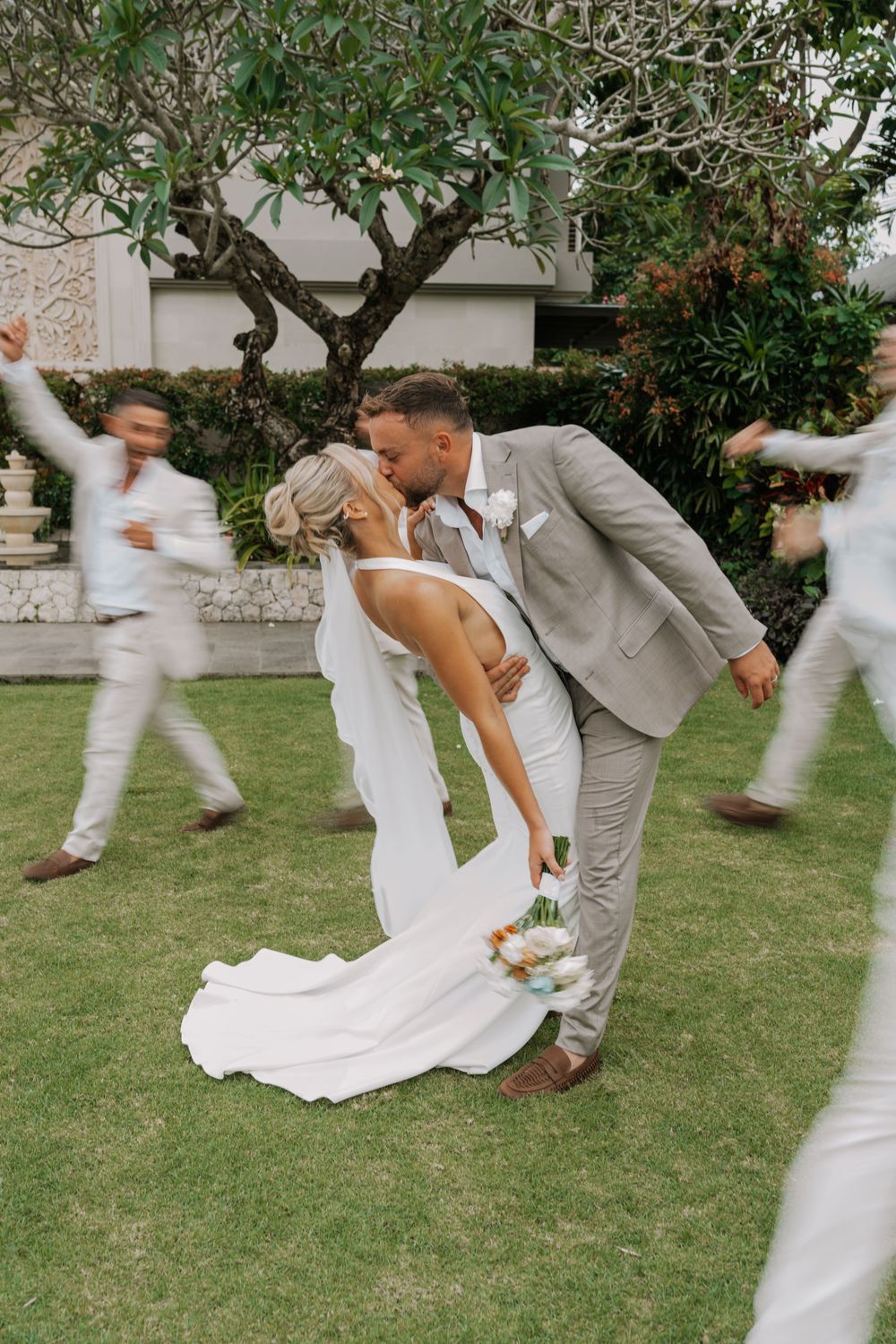 A romantic wedding kiss moment on a grassy lawn with guests in white blurred in motion around the couple.