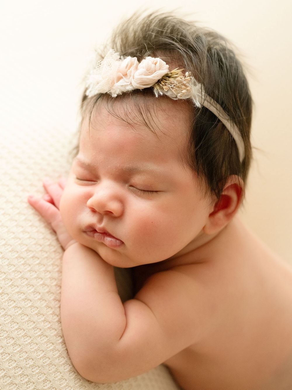 Sleeping newborn baby wearing delicate floral headband on cream textured blanket.