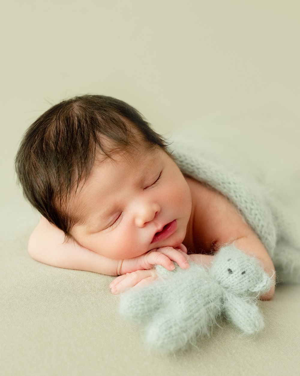 A sleeping newborn in soft white wrap rests peacefully against a pale beige backdrop.