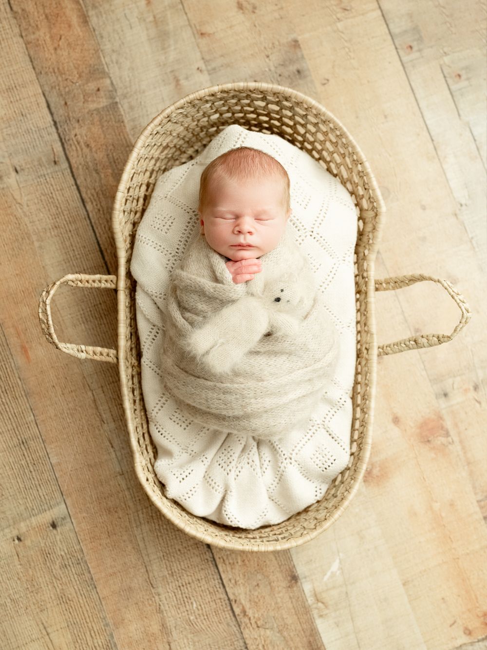 Newborn baby wrapped in soft white blanket sleeps peacefully in round woven basket on rustic wooden floor.