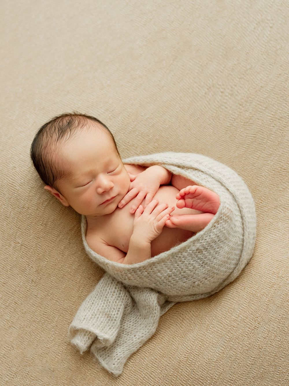 A peaceful newborn sleeping, wrapped in a cozy grey knit blanket on beige fabric background.