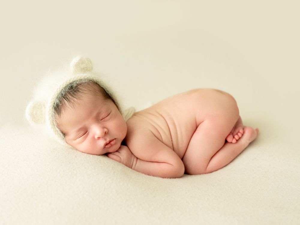 A sleeping newborn baby curled up peacefully on a soft cream-colored backdrop in minimalist photography style.
