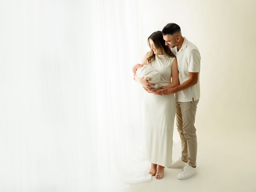 A couple in white outfits embrace lovingly during an intimate photo session against a neutral backdrop.