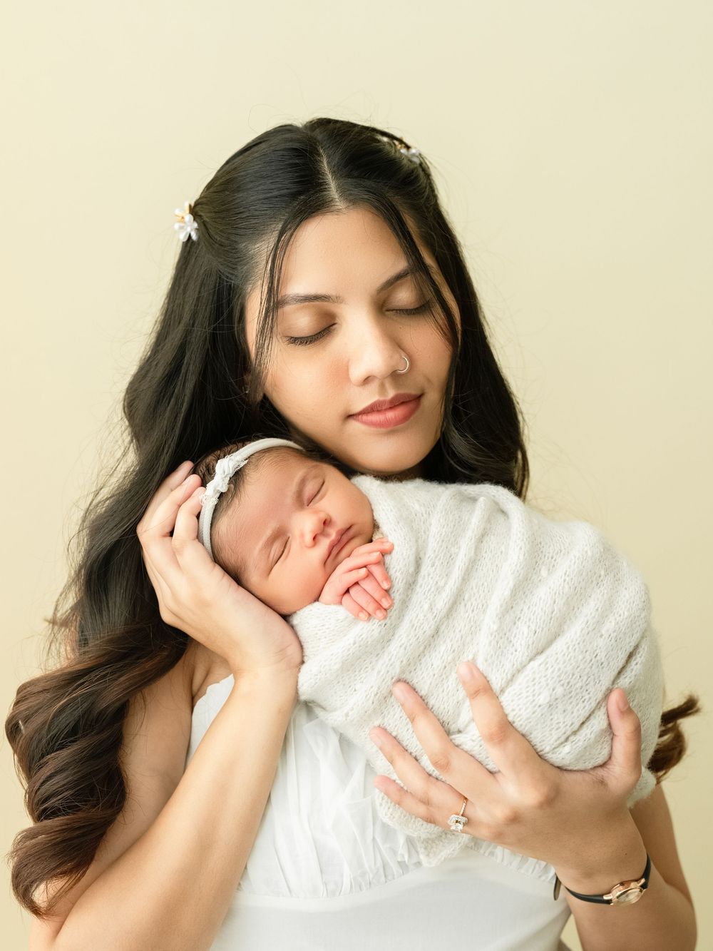 Parent in a white outfit cuddles and bonds with sleeping newborn baby in a white blanket against neutral background.