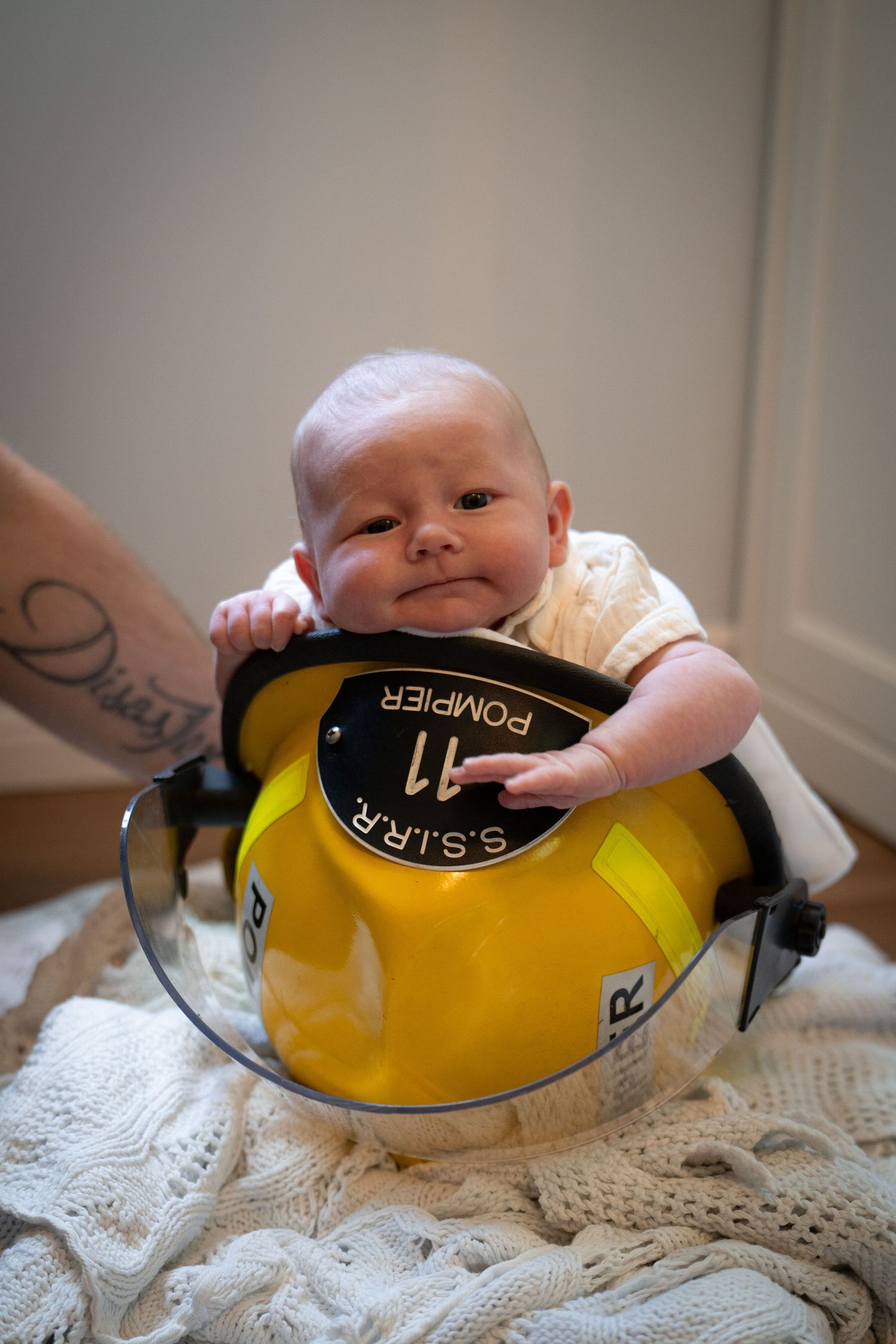 Adorable newborn baby resting inside a yellow POMPIER firefighter helmet on white blanket.