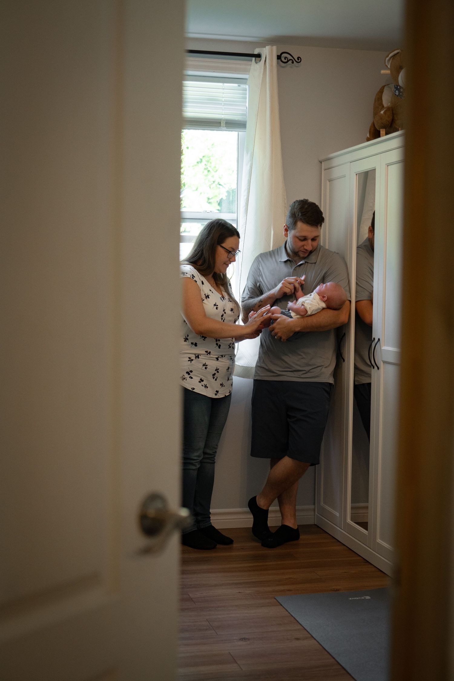 New parents standing in nursery room holding their newborn baby near white wardrobe.