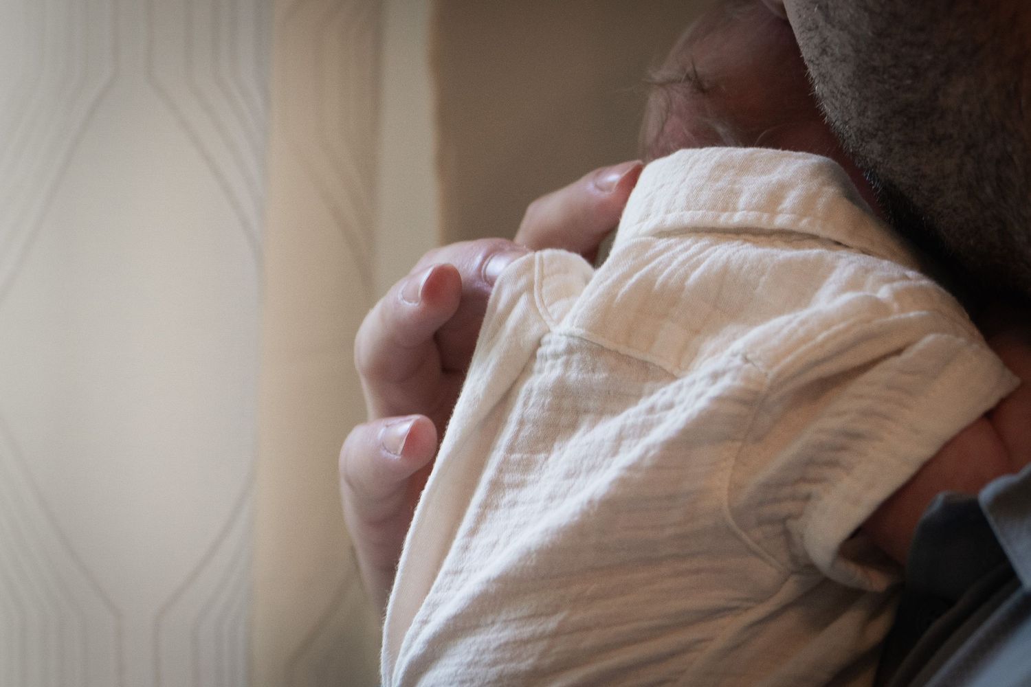 Father tenderly holding newborn baby wrapped in soft muslin swaddle blanket near window.