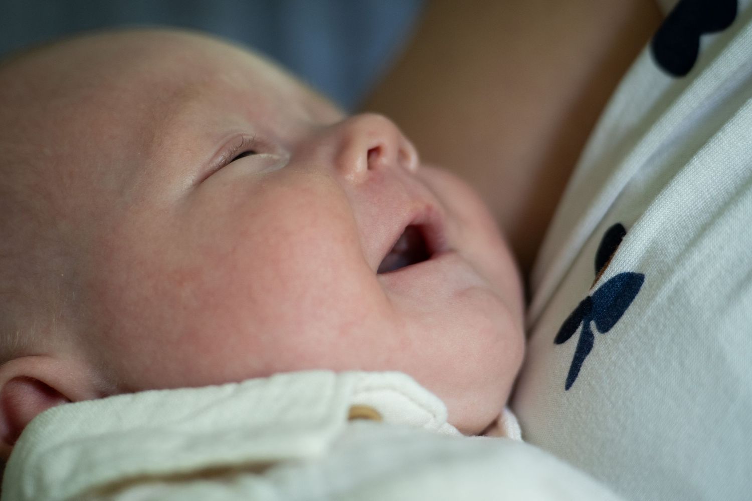 Newborn baby with mouth open being held by mother, close-up of peaceful infant in white blanket.