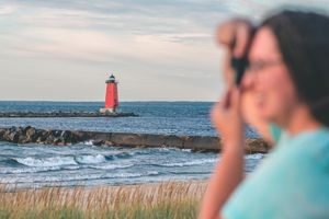 woman takes a photo with a lighthouse and lake behind her