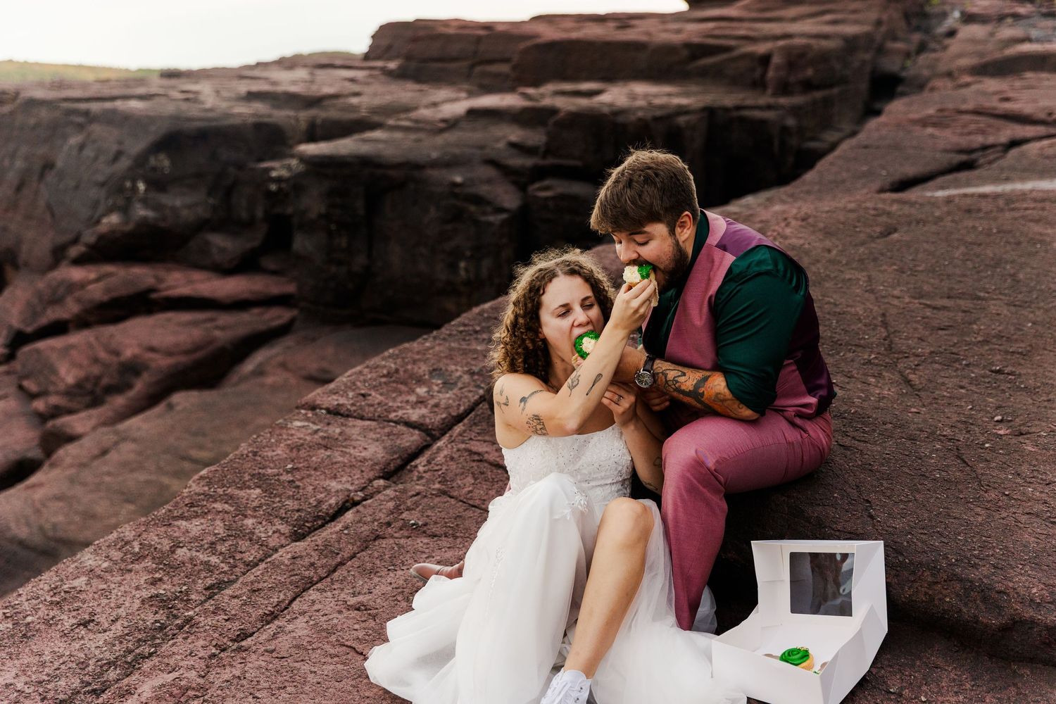 A romantic couple in formal attire shares an intimate moment on red rock formations at sunset.