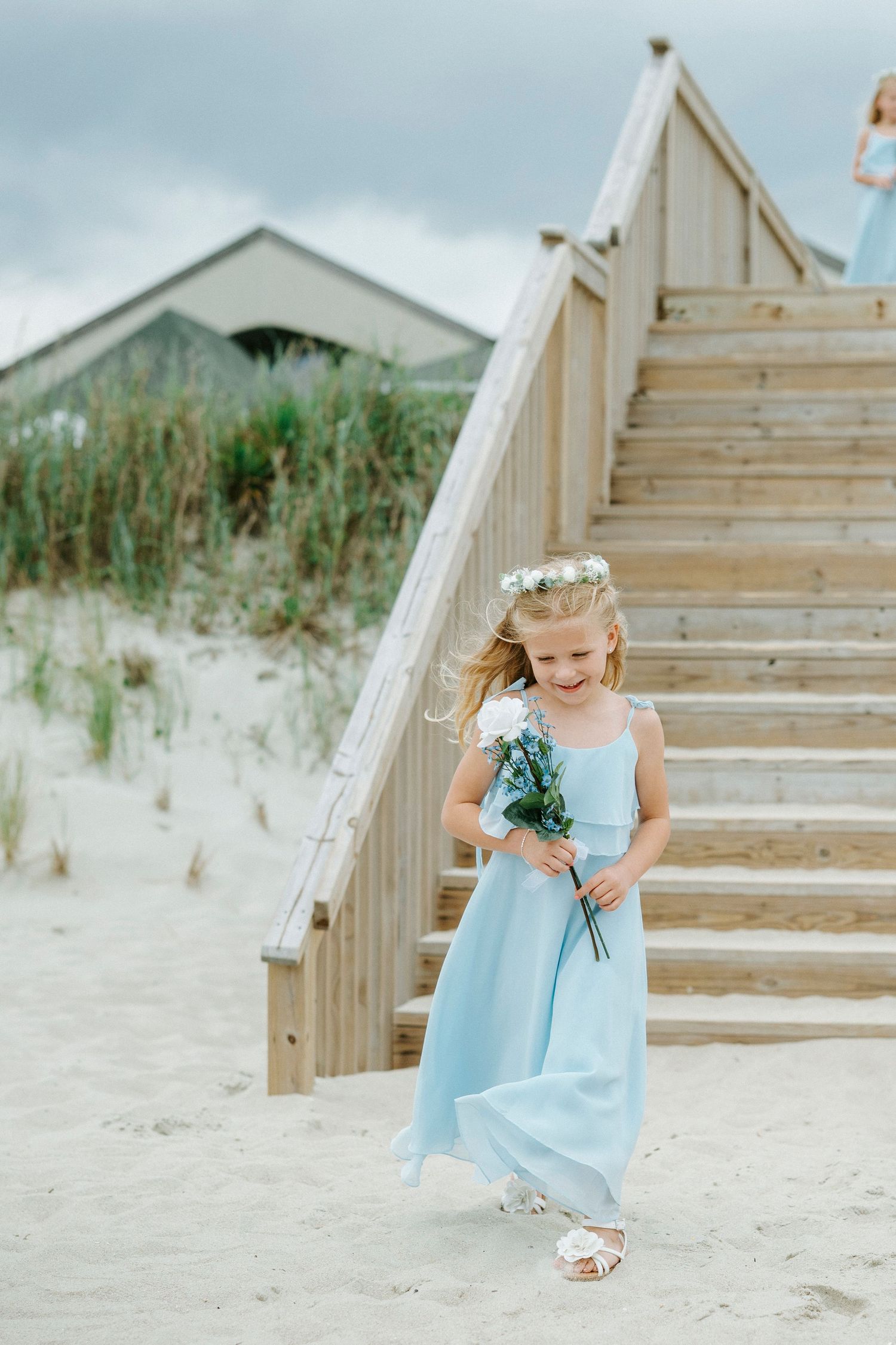A woman in a light blue dress sits gracefully on white wooden beach stairs with sandy dunes in the background.