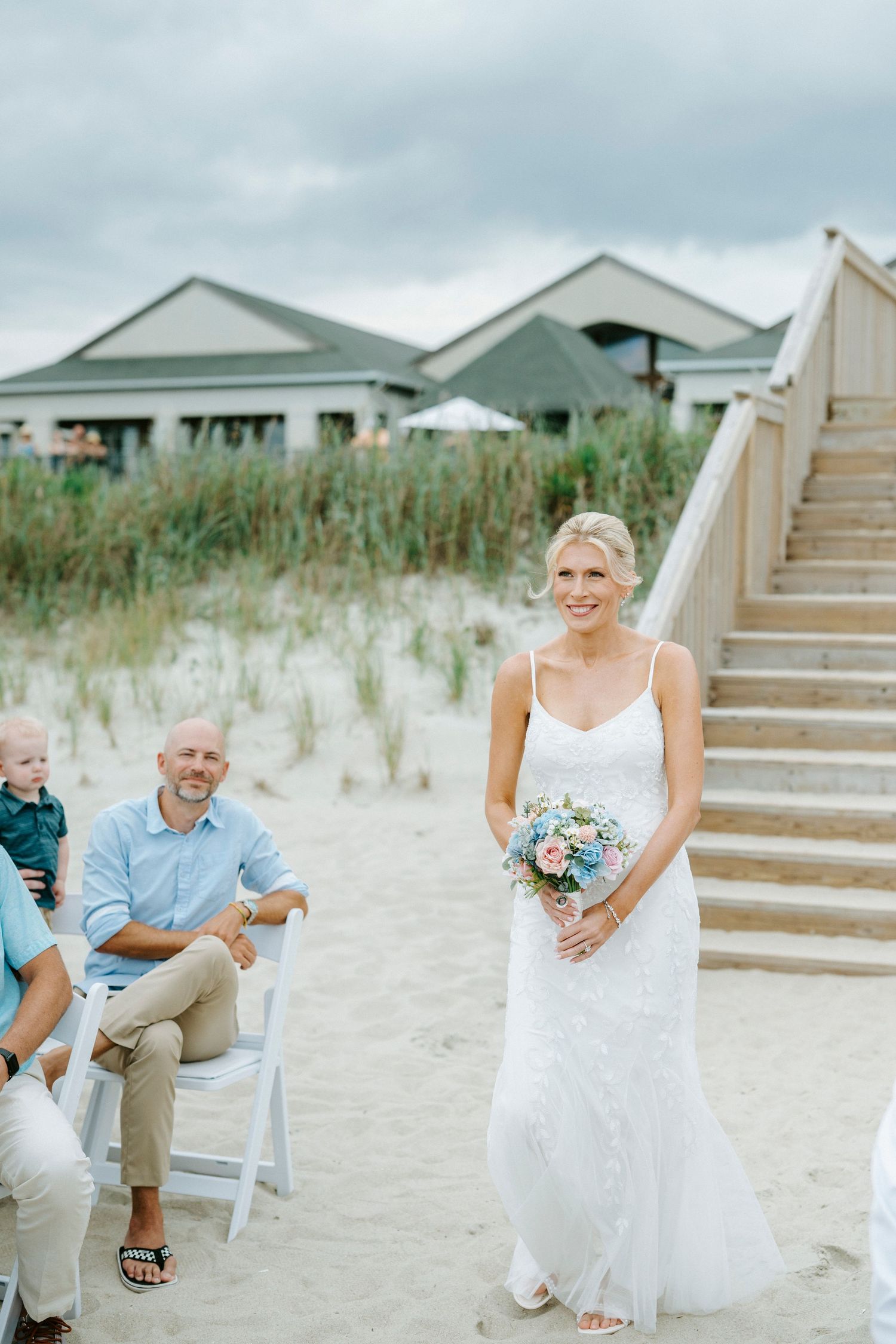 A wedding couple and small child sit together on white chairs along a sandy beach boardwalk near grass dunes.