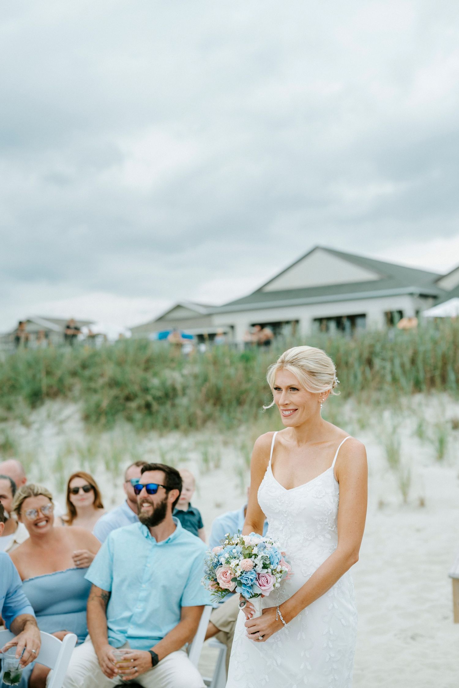 Wedding party seated on white sandy beach with guests wearing light blue attire and coastal homes in background.