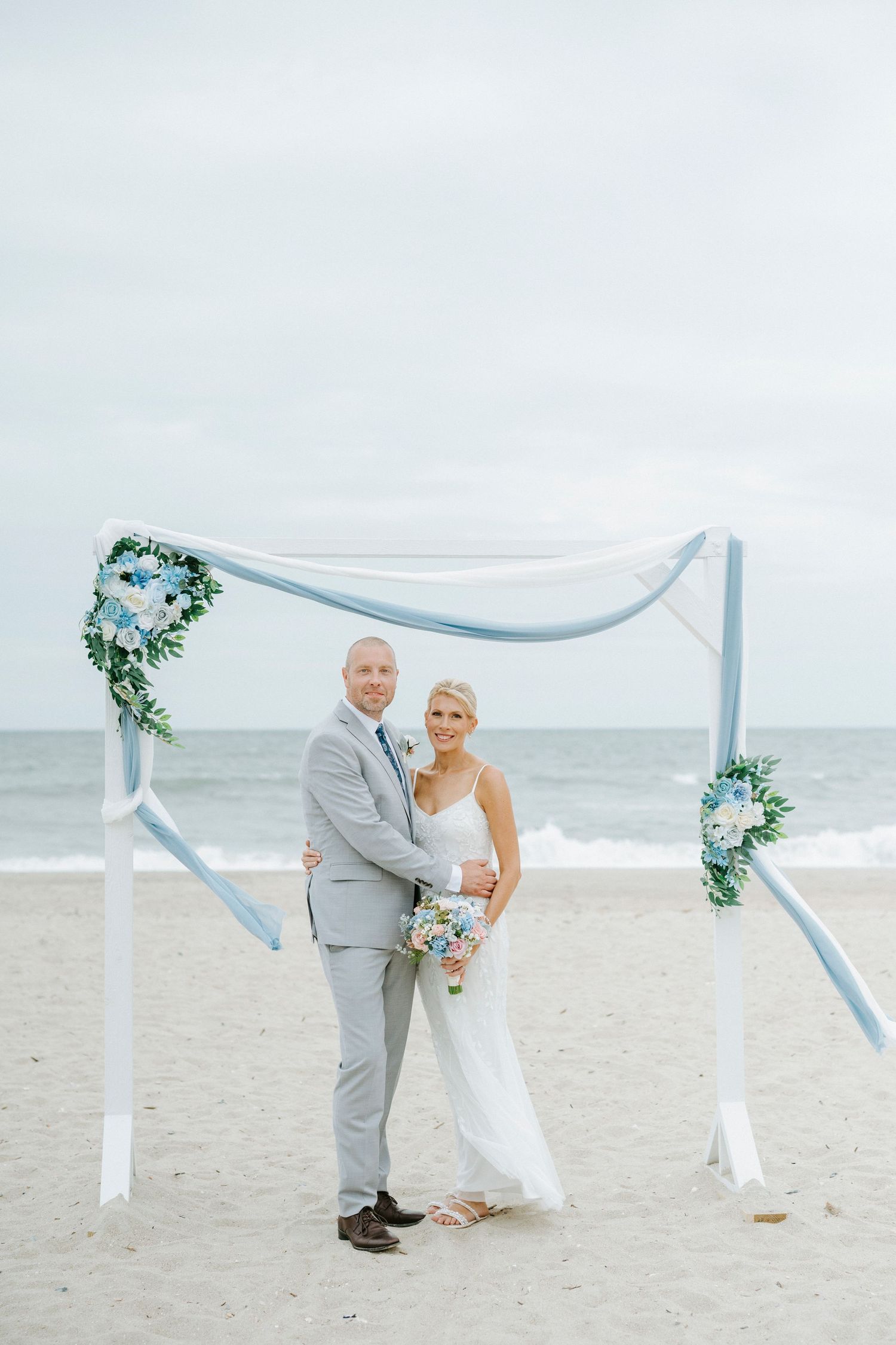 A couple shares a romantic moment under a white draped arch decorated with blue and white flowers on the beach.