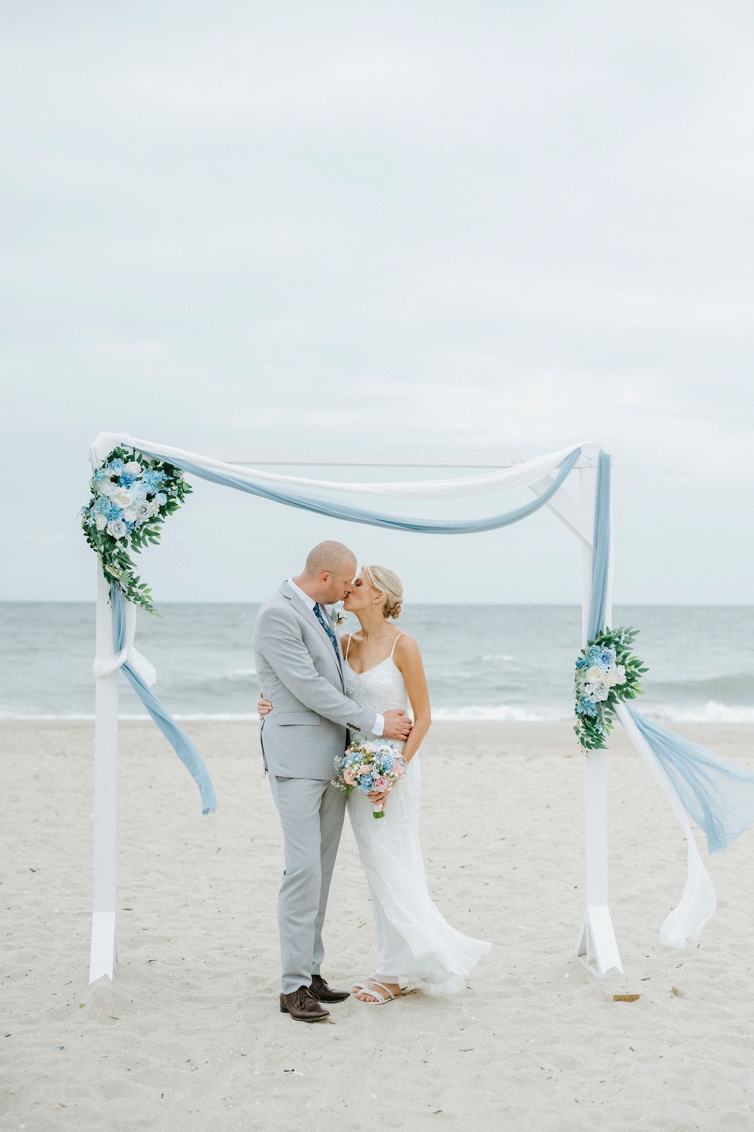 A romantic beach wedding scene with white fabric archway and blue accents decorating the sandy shoreline at dusk.