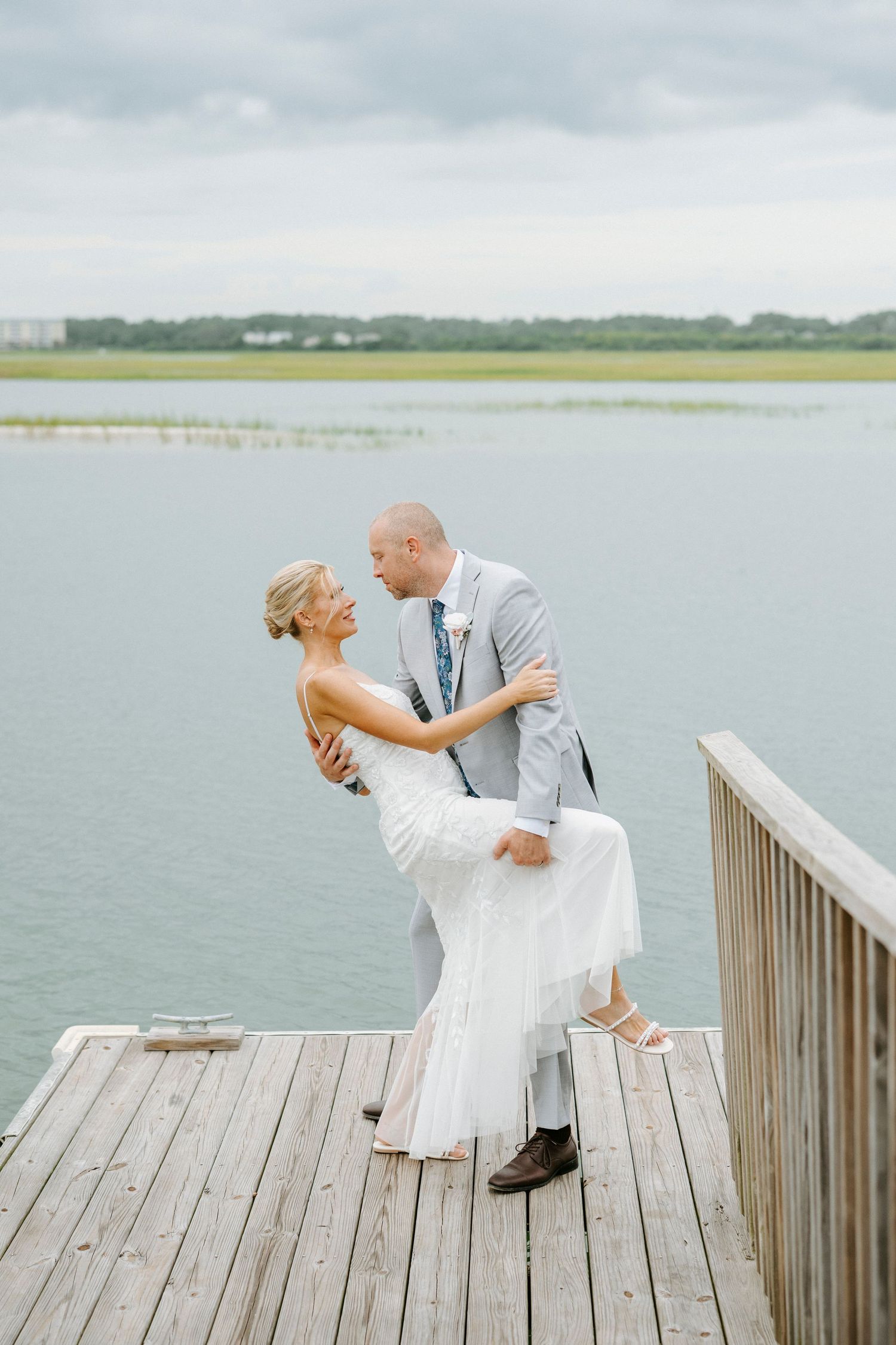 A romantic couple in wedding attire embrace and dance together on a wooden dock overlooking the water.