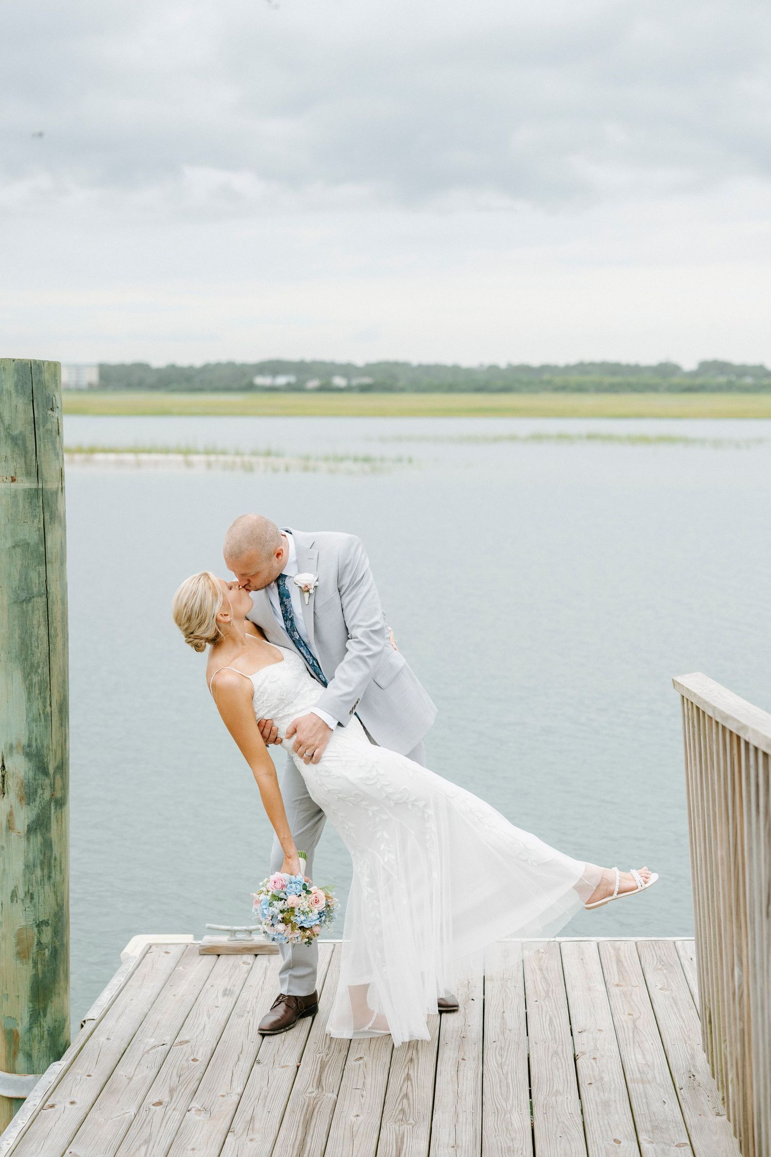 Couple in wedding attire dances playfully on a wooden pier overlooking calm waters.