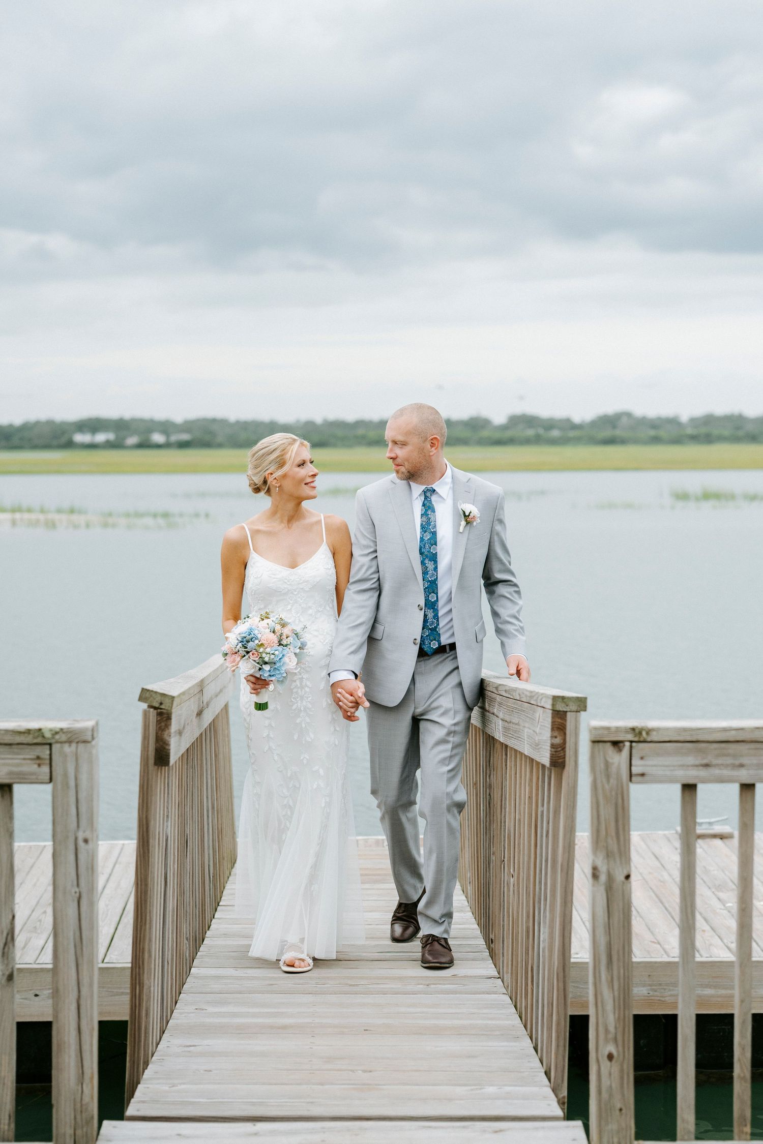 Newlywed couple in white and gray walking together on a wooden pier over tranquil waters on their wedding day.