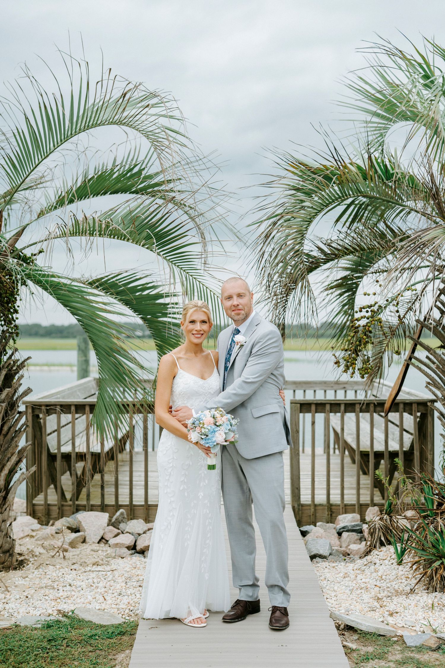 A couple poses on a beach deck surrounded by tropical palm trees and white stones in elegant wedding attire.